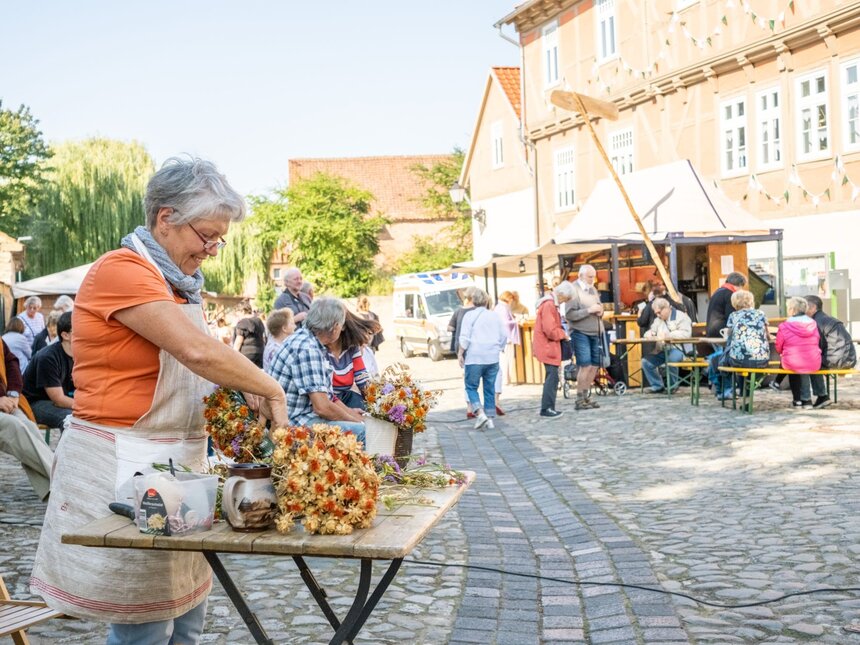 Eine Frau bindet auf einem Kopfsteinpflasterplatz Trockenblumensträuße, dahinter sitzen Gäste an Biertischen vor einem Fachwerkhaus.